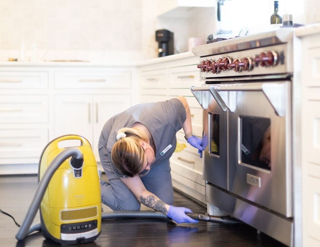 This image depicts a person cleaning the kitchen of a home. They are kneeling on the floor, wearing gloves, and using a vacuum cleaner to clean under the stove. The kitchen appears to be spacious and modern, with white cabinets and a stainless steel stove. The person is likely part of a professional cleaning service, providing regular house cleaning or residential cleaning services to the homeowner.