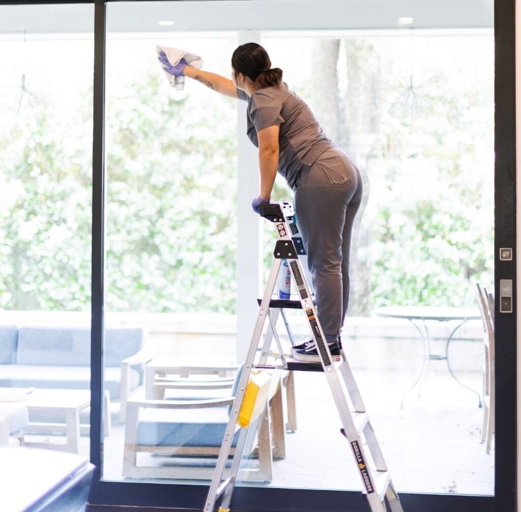 A woman is standing on a stepladder, cleaning a large window in a home. She's wearing gloves and holding a cleaning cloth. The window has a view of a patio with outdoor furniture and greenery. The woman is likely part of a professional cleaning service, providing regular house cleaning or residential cleaning services to the homeowner.