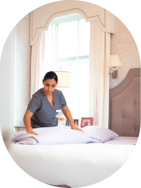 The image shows a person, likely a housekeeper or cleaning professional, making a bed in a well-lit, elegantly furnished bedroom. The person is wearing a grey uniform, which suggests they are working in a professional cleaning role. The bed has a soft headboard, and the room features light-colored walls and curtains, with decorative elements like framed pictures on a nightstand. The scene reflects cleanliness and attention to detail, potentially representing the services offered by a cleaning company such as Vella Clean in Austin.