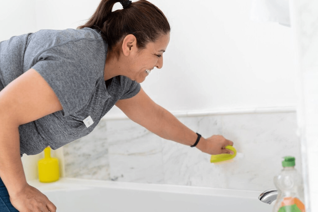 The image shows a smiling cleaning professional in a grey uniform, scrubbing the side of a bathtub with a yellow sponge. 