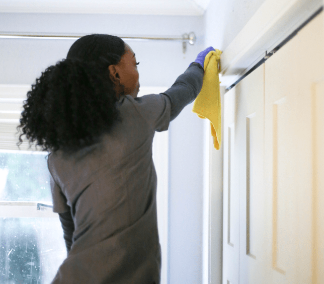 A professional cleaner in a grey uniform reaching up to dust window frames and door trim
providing thorough residential cleaning services at a client's home