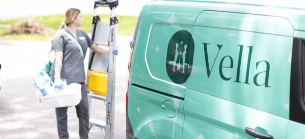 A housekeeping professional stands by a teal van labeled "Vella," carrying cleaning supplies and a ladder, ready for service.