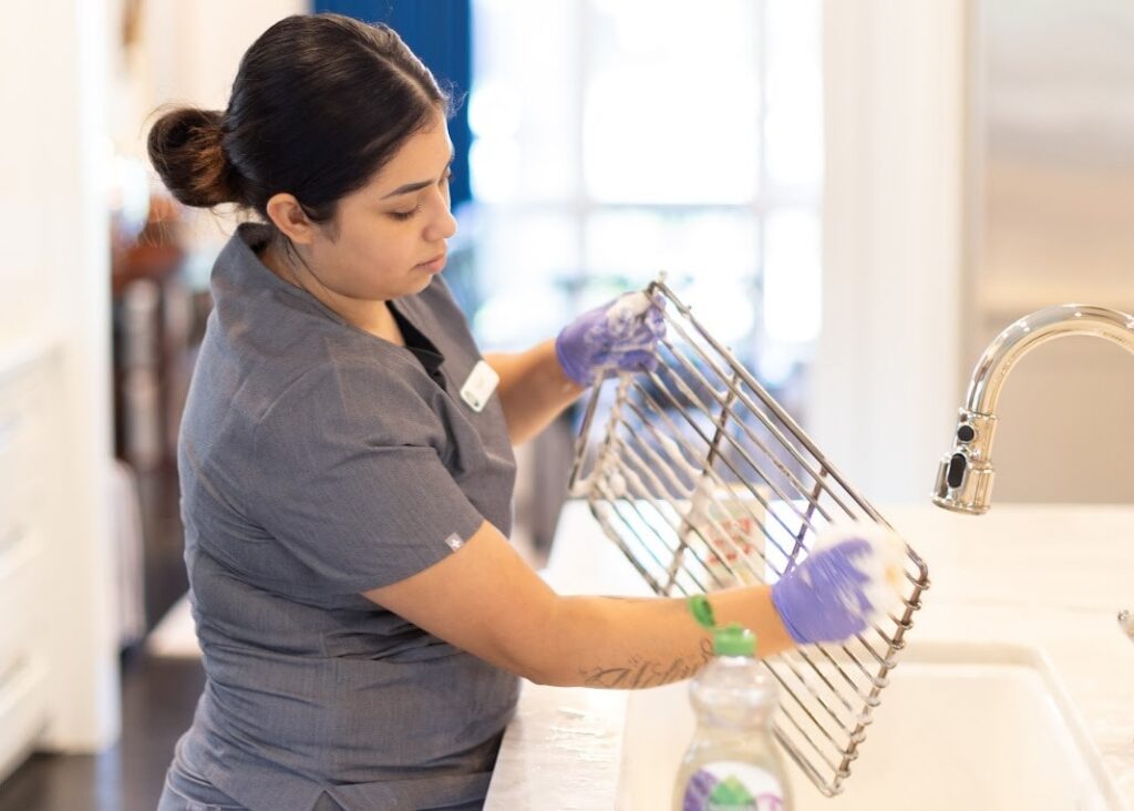 A professional cleaner wearing gloves carefully scrubs a metal rack over the sink, demonstrating how to deep clean your kitchen for a spotless result.