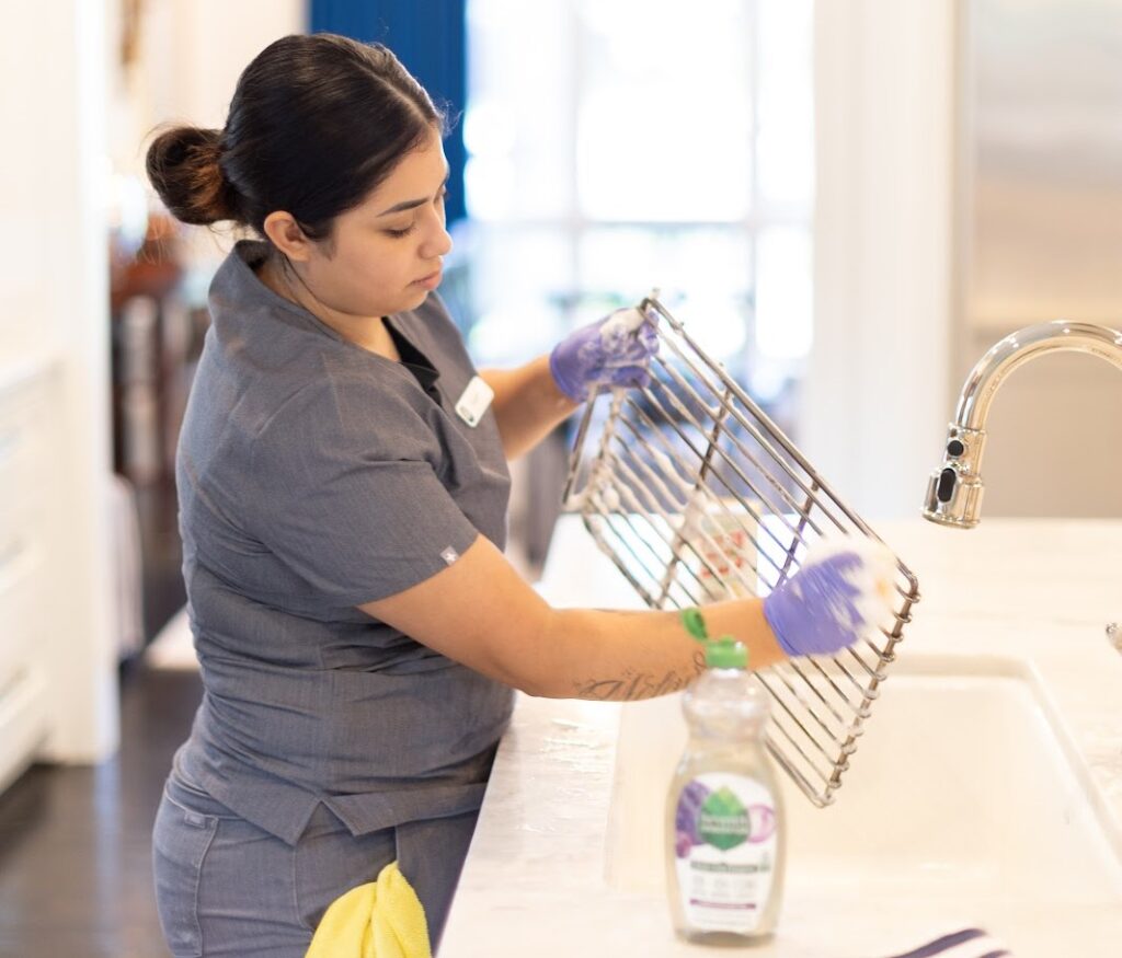 Person performing regular home cleaning by scrubbing a metal grill rack at a sink, wearing a grey uniform and purple gloves, with cleaning supplies on the counter.