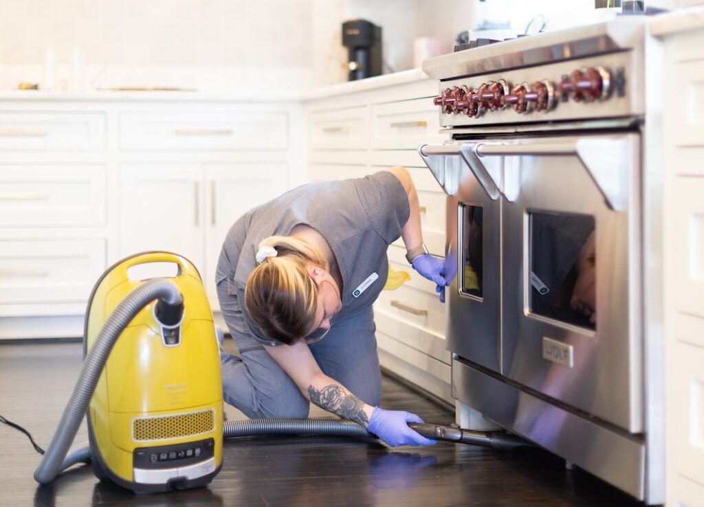 A professional cleaner using a vacuum to clean under a kitchen stove in a modern kitchen; remember to clean your sink thoroughly for a spotless kitchen.