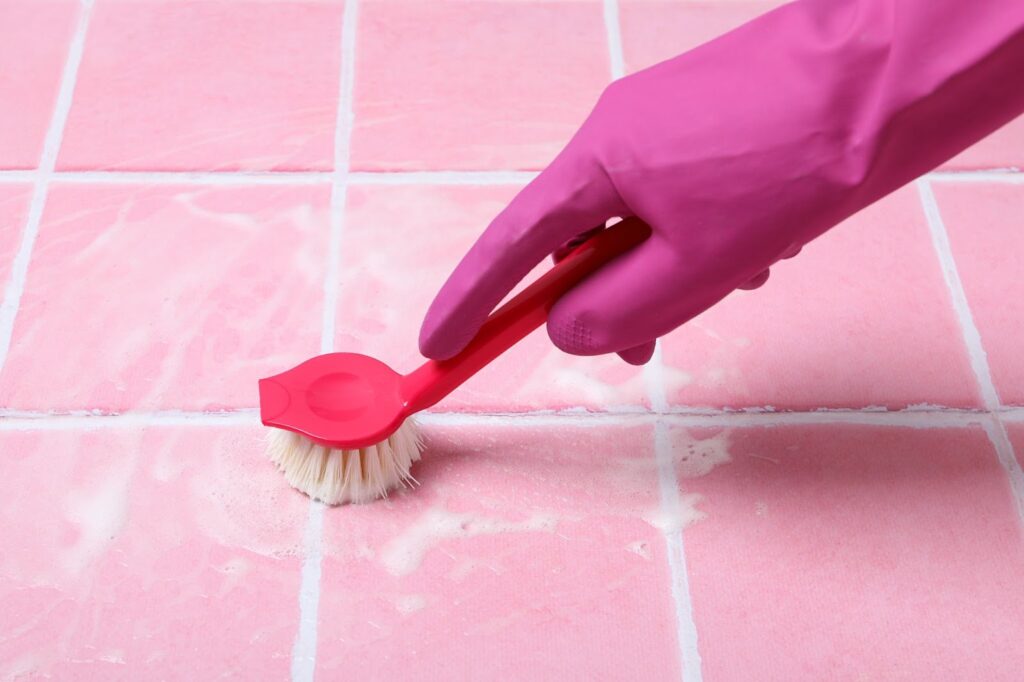 A gloved hand holding a pink scrub brush on a pink tiled floor.