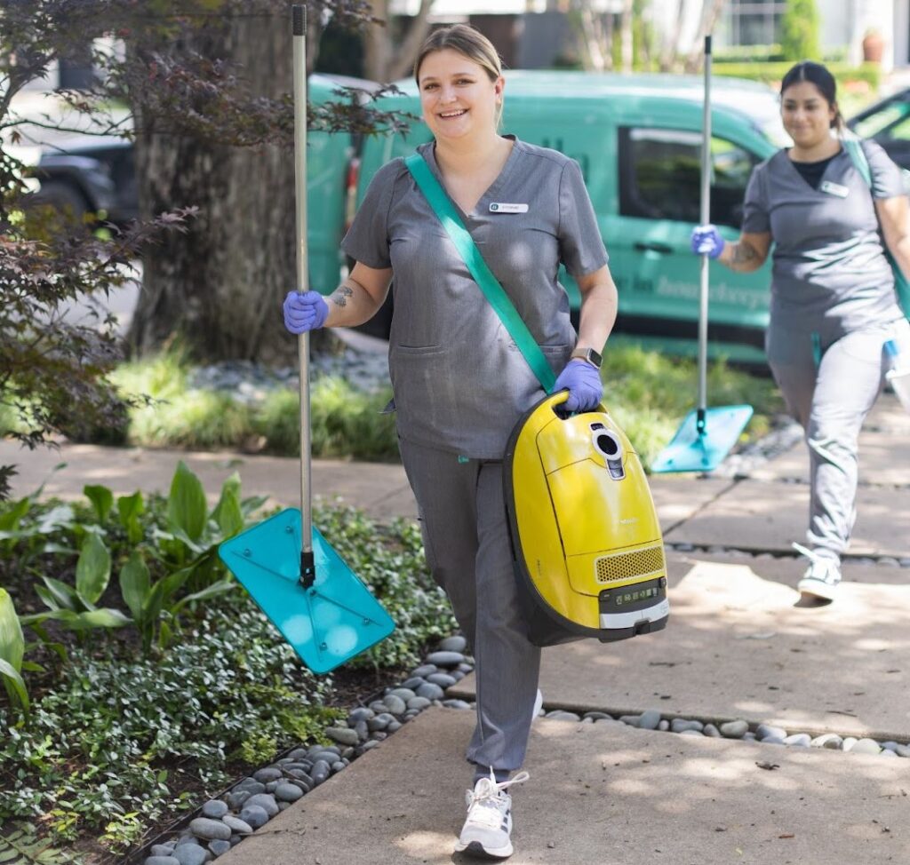 A smiling woman in a gray uniform carrying a yellow vacuum cleaner 