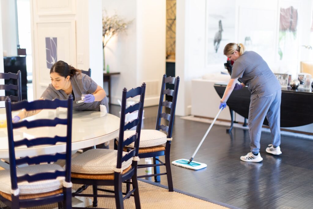 Two cleaning professionals tidying up a modern dining and living space, one wiping a table and the other mopping the floor.