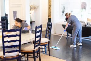 Two cleaning professionals housekeeping up a modern dining and living space, one wiping a table and the other mopping the floor.