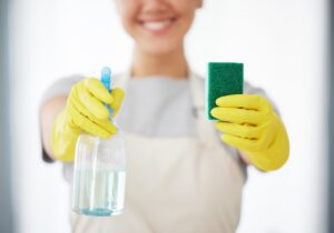 Person holding a green sponge and a spray bottle, wearing yellow gloves and a light apron, prepared for regular home cleaning.