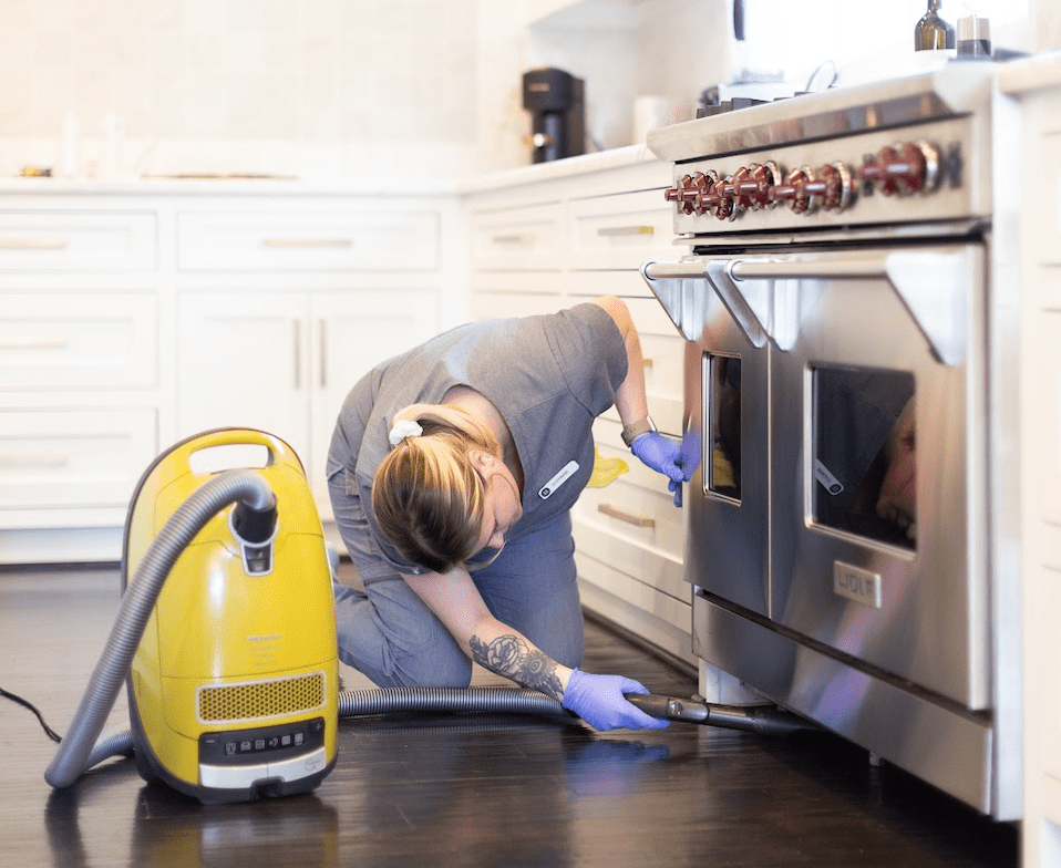 A cleaner uses a yellow vacuum to deep clean under a kitchen appliance, showcasing deep cleaning services in Austin.