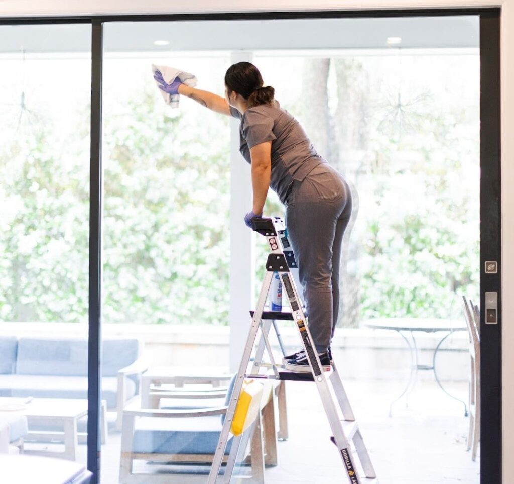 A professional cleaner wiping a large glass window while standing on a ladder.