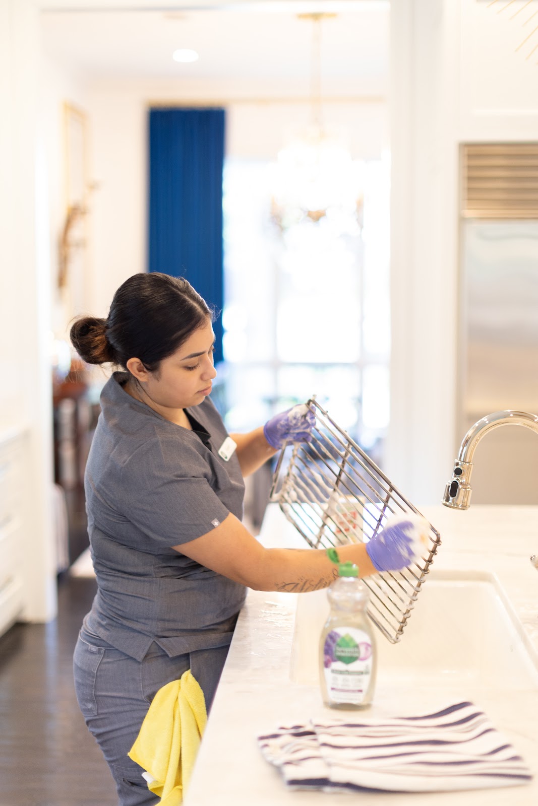 Housekeeping staff member cleaning a metal kitchen rack at a sink in a well-lit modern kitchen, wearing gloves and a uniform.