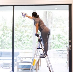 Professional cleaner performing specialty cleaning on large glass doors, standing on a step ladder and ensuring a spotless finish, with a serene outdoor view in the background.