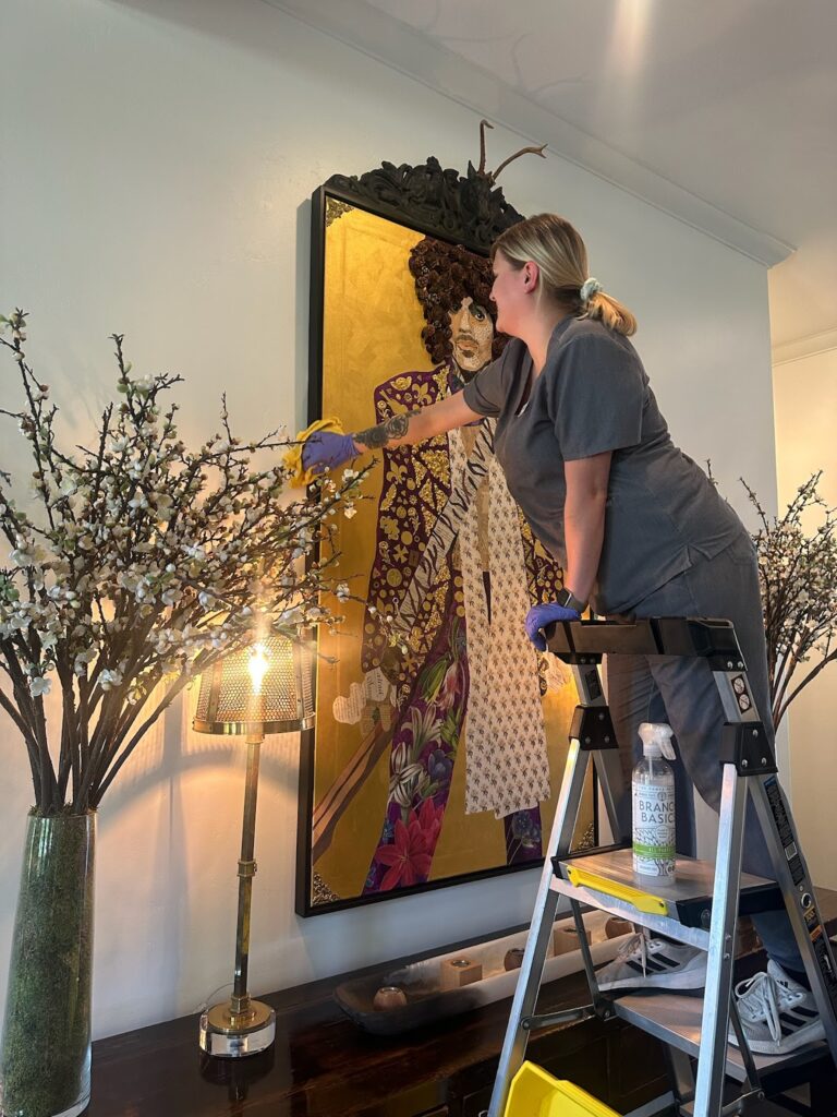 Professional cleaner performing specialty cleaning, carefully dusting a vibrant wall art piece and decor while standing on a step ladder, surrounded by elegant interior elements.