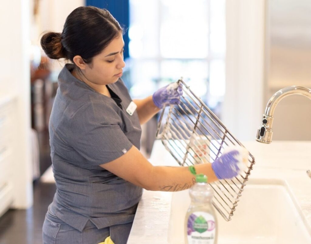 A professional cleaner scrubbing a metal rack over a sink with soap and gloves.