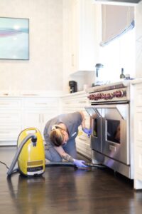 Vella cleaner vacuuming under a kitchen stove with a Milie vacuum in Fort Worth, Texas.