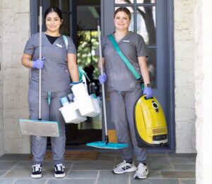 Vella’s cleaning professionals are standing in front of the main door of a house with cleaning products, a mop, and a vacuum cleaner.