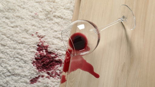 Red wine spilled from a glass onto a light wood floor and white carpet in NYC home.