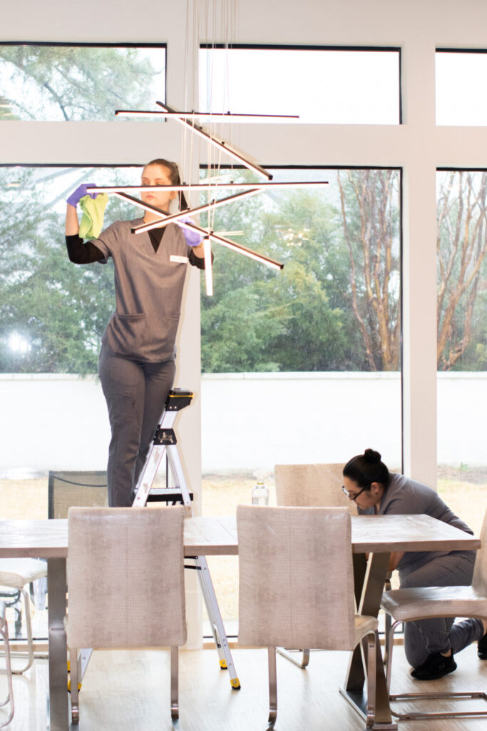 Two cleaning professionals tidying a modern dining room, one wiping a light fixture, NYC.