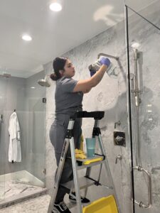 Vella’s professional house cleaner is cleaning the shower inside a bathroom while standing on a ladder.