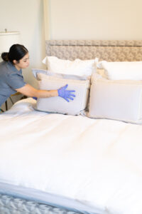 A Vella cleaner in uniform adjusts pillows on a neat bed, ensuring a welcoming space.