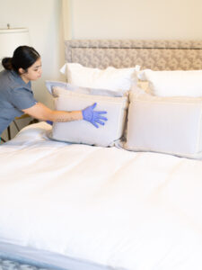 A Vella cleaner in uniform adjusts pillows on a neat bed, ensuring a welcoming space.