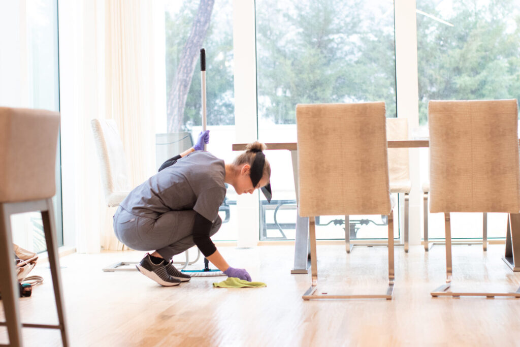 A housekeeper in gray uniform cleaning a wooden floor in a dining room, using a green cloth near a table and chairs.