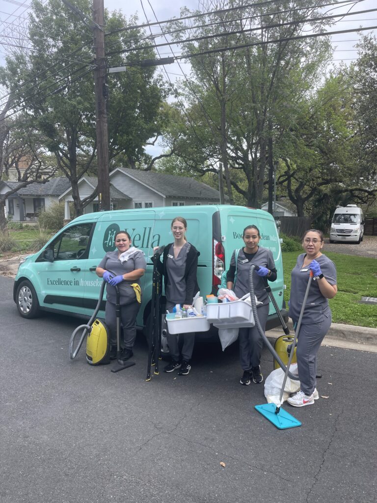 Four Vella professional house cleaners stand near a van, smiling, and holding cleaning equipment.