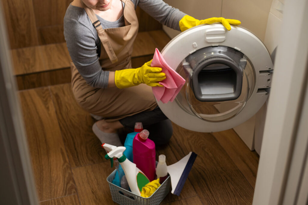 A Vella cleaner in gloves wipes a washing machine door with a cloth, surrounded by eco-friendly cleaning supplies.
