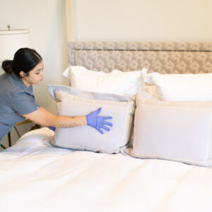 A Vella cleaner in uniform adjusts pillows on a neat bed, ensuring a welcoming space.