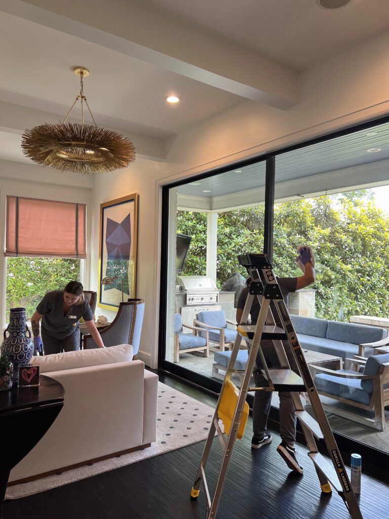 Two cleaning staff members are tidying a modern living room and washing large glass patio doors.