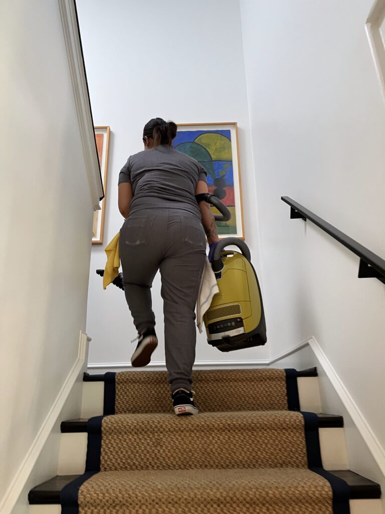 A Vella cleaner carrying a vacuum up the stairs while preparing for a detailed home cleaning session.