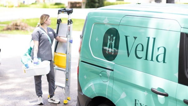 A professional cleaner unloading supplies from a Vella-branded van parked outside a residential house in Dallas.
