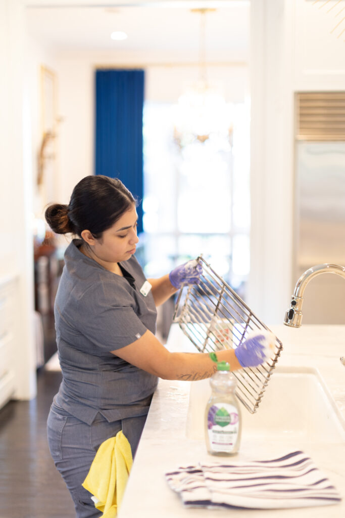 Cleaner scrubbing a metal kitchen rack with eco-friendly soap at a sink, wearing gloves and a uniform.